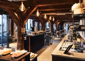 En rymlig bufférestaurang i rustik stil med synliga träbjälkar och varm belysning. Borden är dukade med en mängd olika rätter, och stora fönster tillåter naturligt ljus att fylla utrymmet.