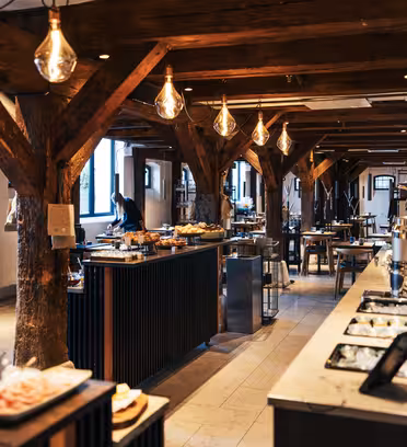 En rymlig bufférestaurang i rustik stil med synliga träbjälkar och varm belysning. Borden är dukade med en mängd olika rätter, och stora fönster tillåter naturligt ljus att fylla utrymmet.