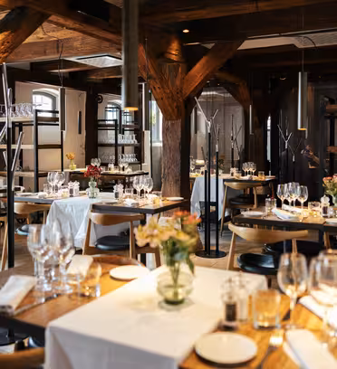 En mysig restauranginredning med träbjälkar och varmt upplysta bord. Vita dukar och snyggt arrangerade kuvert syns, tillsammans med blommor och glas på borden. Atmosfären är elegant och inbjudande.