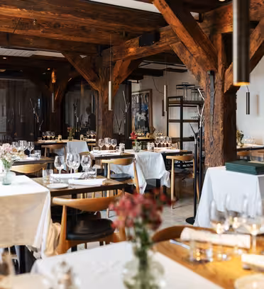 En mysig restauranginredning med träbjälkar och bord för middagar. Borden är täckta med vita dukar, prydda med glas och blomsterarrangemang. Mjuk belysning skapar en varm och inbjudande atmosfär.