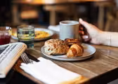 En person som håller i en mugg sitter vid ett träbord med en tallrik med bakverk, inklusive en croissant och en frörulle. I närheten finns en tidning, ett glas juice och ett annat glas. Bakgrunden är mjukt suddig.