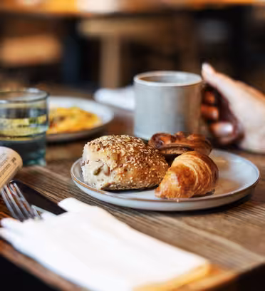 En person som håller i en mugg sitter vid ett träbord med en tallrik med bakverk, inklusive en croissant och en frörulle. I närheten finns en tidning, ett glas juice och ett annat glas. Bakgrunden är mjukt suddig.