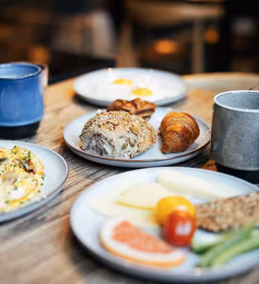 Ett dukat frukostbord med tallrikar som innehåller en omelett med örter, en fralla med frö, en croissant, ostskivor, en körsbärstomat, gurkstavar och en grapefruktskiva. Det finns även koppar kaffe och glas vatten.