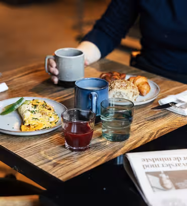 En person som sitter vid ett träbord med en tallrik med omelett och grönsaker, en annan med bröd och en liten croissant. Det finns två muggar, ett glas röd juice och ett glas vatten. En tidning är delvis synlig på bordet.