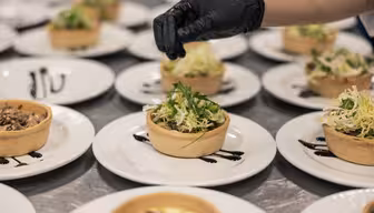 En kock som bär en svart handske garnerar en rad små salta tårtor. Tårtorna är fyllda med grönt och andra ingredienser, placerade på vita tallrikar med en klick mörk sås, arrangerade på en yta av rostfritt stål.