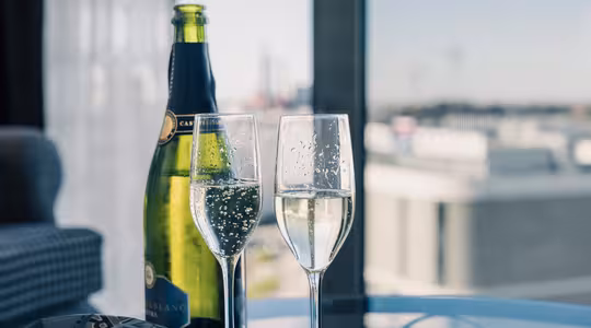 Två fyllda champagneflöjter och en flaska mousserande vin sitter på ett bord vid ett fönster. Solljus framhäver bubblorna i glasen. Stadsbilden utanför är något suddig, vilket skapar en lugn och festlig atmosfär.