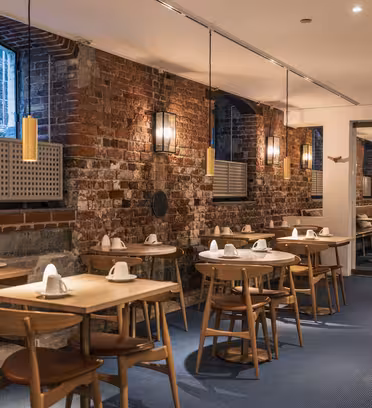 Mysig restaurang med exponerade tegelväggar, träbord och stolar och mjuk omgivningsbelysning. Borden är dukade med vita servetter. Stora fönster släpper in naturligt ljus och en spegel reflekterar utrymmet och skapar en öppen känsla.