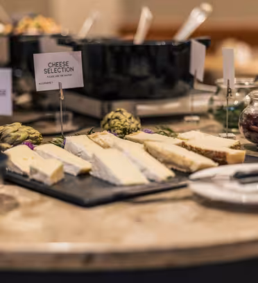 En ostbricka på ett marmorbord innehåller olika ostar med etiketter, kronärtskockor och en burk konserver. I bakgrunden finns svarta serveringsgrytor. Tallrikar och bestick finns i närheten.