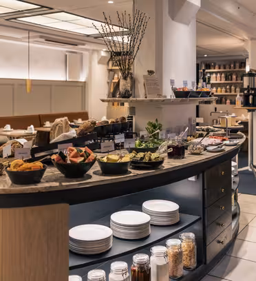 En buffé i en elegant matsal med olika frukter, bakverk och frukostprodukter. Tallrikar och burkar med kryddor står uppställda på en böjd disk. Utrymmet har mysiga sittplatser med neutrala färger och varm belysning.