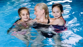 En kvinna ler när hon står i en pool och håller två unga flickor i baddräkter. Det klarblå vattnet omger dem när de njuter av sin tid tillsammans.