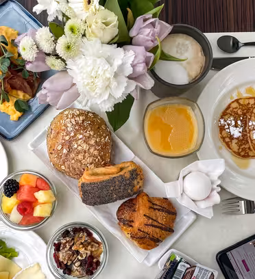 En frukost pålägg med äggröra och bacon, pannkakor med socker, diverse bröd, bakverk, färsk frukt, yoghurt med nötter och apelsinjuice. En smartphone och latte står på bordet bredvid en vas med vita och lila blommor.