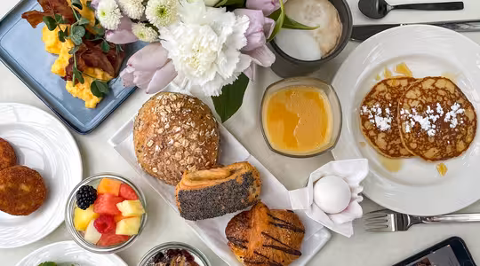 En frukost pålägg med äggröra och bacon, pannkakor med socker, diverse bröd, bakverk, färsk frukt, yoghurt med nötter och apelsinjuice. En smartphone och latte står på bordet bredvid en vas med vita och lila blommor.