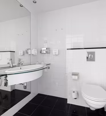 Modernt badrum med vita kaklade väggar och svart klinkergolv. Har en väggmonterad toalett, stor spegel, handfat i glas med handduk under och ett badkar med duschvägg till höger.