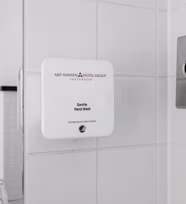 En dispenser för vit flytande tvål märkt "Gentle Hand Wash" är monterad på en vit kaklad vägg. En mjukpappershållare i rostfritt stål är delvis synlig på höger sida. En del av en glasyta ses till vänster.