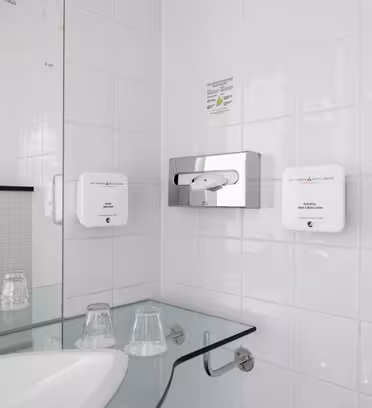 Ett modernt badrum med vita kaklade väggar har en väggmonterad hårtork mellan två tvålautomater. En glashylla rymmer två plastmuggar nära ett vitt handfat. Området är rent och minimalistiskt, med en dusch synlig till vänster.