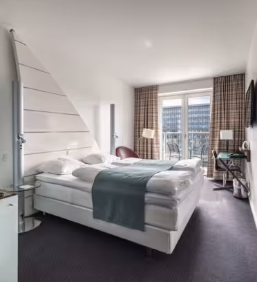 Ett modernt hotellrum med stor säng, blågrön löpare och vita sängkläder. Rummet har ett träskåp, väggmonterad TV och ett litet runt bord med två stolar. Ett fönster med rutiga gardiner ger utsikt över närliggande byggnader.