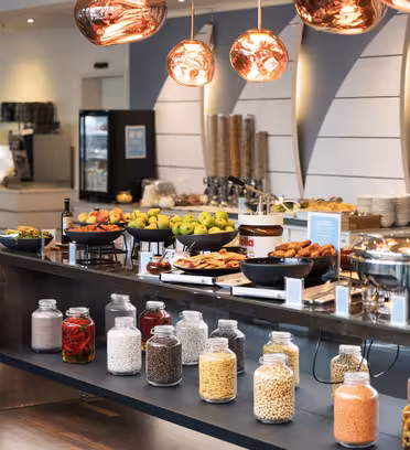En bufféuppsättning med burkar med olika spannmål och spannmål, fruktskålar, bakverk och skavrätter. Kopparhängande lampor hänger ovanför, och bakgrunden inkluderar en kaffemaskin och ett kylskåp.