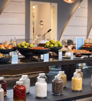 Bufféuppsättning med diverse frukter, bakverk och bröd på en träbänk. Nedanför rymmer hyllorna burkar med olika spannmål och pasta. Miljön är modern med fokus på färska råvaror och en elegant design.