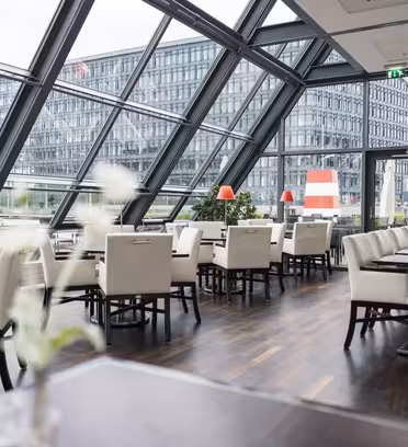 Modern restauranginteriör med eleganta vita stolar och mörka träbord. Stora snedställda fönster ger utsikt över en modern kontorsbyggnad. Röda lampor och minimala blommutter lägger till accenter till den moderna designen.