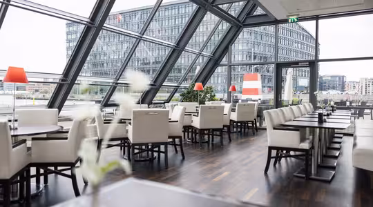 Modern restauranginteriör med eleganta vita stolar och mörka träbord. Stora snedställda fönster ger utsikt över en modern kontorsbyggnad. Röda lampor och minimala blommutter lägger till accenter till den moderna designen.