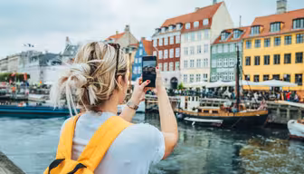 En person med blont hår och en gul ryggsäck tar ett foto med en smartphone. De står inför färgglada byggnader och båtar längs en kanal, under en molnig himmel. Scenen är livlig med människor och vatten som speglar omgivningen.