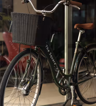 En grön cykel med en svart vävd korg på framsidan står parkerad innanför en glasentré. Cykeln har bruna handtag och ett brunt säte. En skinande grön skulptur syns i bakgrunden.