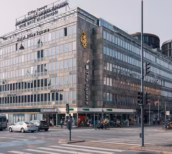 Gatuvy av en upptagen korsning i en stad. En stor hörnbyggnad med skyltning till "Hotel The Square" dominerar scenen. Det finns olika butiker på bottenvåningen, bilar, cyklar och fotgängare syns på gatan.