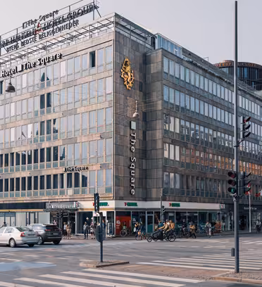 Gatuvy av en upptagen korsning i en stad. En stor hörnbyggnad med skyltning till "Hotel The Square" dominerar scenen. Det finns olika butiker på bottenvåningen, bilar, cyklar och fotgängare syns på gatan.
