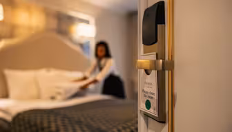 En hotellrumsdörr med skylten "Please clean the room" står på glänt. I bakgrunden bäddar en hushållerska sängen och rummet har elegant inredning med varm belysning.