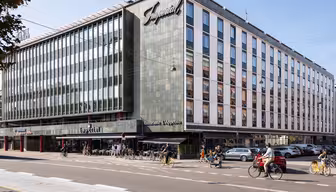 Stor byggnad med modern fasad och en skylt med texten Imperial. Bottenvåningen rymmer en restaurang med uteservering. Folk cyklar och går längs gatan. Flera parkerade bilar syns framför. Klarblå himmel i bakgrunden.