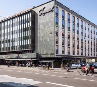 Stor byggnad med modern fasad och en skylt med texten Imperial. Bottenvåningen rymmer en restaurang med uteservering. Folk cyklar och går längs gatan. Flera parkerade bilar syns framför. Klarblå himmel i bakgrunden.