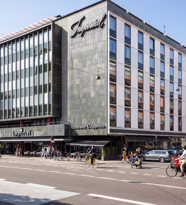 Stor byggnad med modern fasad och en skylt med texten Imperial. Bottenvåningen rymmer en restaurang med uteservering. Folk cyklar och går längs gatan. Flera parkerade bilar syns framför. Klarblå himmel i bakgrunden.