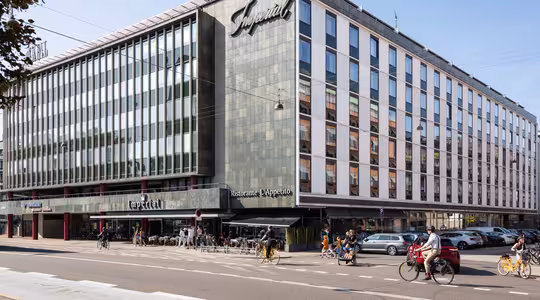 Stor byggnad med modern fasad och en skylt med texten Imperial. Bottenvåningen rymmer en restaurang med uteservering. Folk cyklar och går längs gatan. Flera parkerade bilar syns framför. Klarblå himmel i bakgrunden.