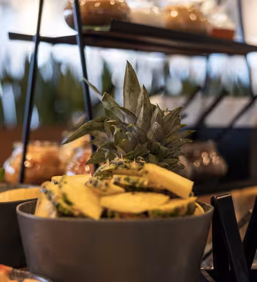 En buffé med en skål fylld med ananasskivor, toppad med en hel ananas som står upprätt. Runt ananasen finns krukväxter och andra rätter, med suddig grönska i bakgrunden.