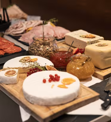 Ett gourmetost- och charkuteripålägg med brie, runda och fyrkantiga ostar, charkuterier, nötter i burk, gula såser, garnityr och kex. Knivar och serveringsredskap placeras i närheten, placerade på träskivor.