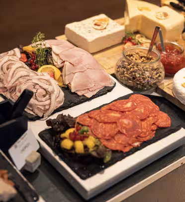 En mängd olika delikatesser och ostar arrangerade på ett buffébord. Tallrikar innehåller skivad skinka, salami och diverse ostar. Burkar med kryddor och garnering som frukt och örter är också synliga. Etiketter och serveringsredskap finns.