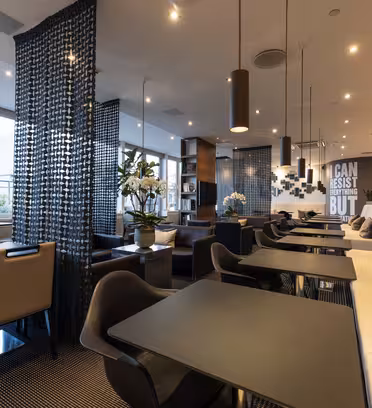 Modern caféinteriör med svarta och beiga möbler. Svarta bord är ihopkopplade med svarta och bruna stolar. Vita bänkar kantar en vägg. Det finns dekorativa skiljeväggar och krukväxter, med stora fönster som släpper in naturligt ljus.