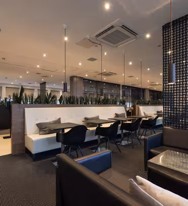 Modern restauranginteriör med eleganta svarta stolar, vita bås och bord med glas. Dekorativa växtavdelare och geometriskt mönstrade väggar skapar en stilfull atmosfär. Takspotlights och stora fönster ger gott om ljus.