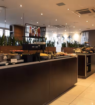 Bufféarrangemang i en modern restaurang med en mängd olika rätter, inklusive bröd, frukt och drycker. Miljön har en elegant design med trä- och metallelement, inomhusväxter och takbelysning, vilket skapar en välkomnande atmosfär.