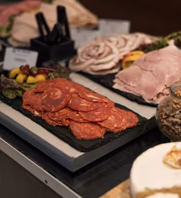 En närbild av en delikatessdisk som visar skivat chark, inklusive chorizo och skinka, tillsammans med en burk valnötter och diverse ost. I bakgrunden syns annat kött och garnityr på svarta serveringsbrickor.