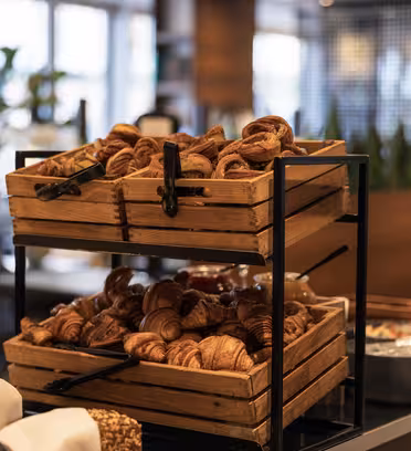Två trälådor fyllda med croissanter är staplade på ett metallställ i en buffémiljö. Bakgrunden är mjukt suddig och visar andra bufféartiklar och en växt, vilket skapar en mysig och inbjudande atmosfär.