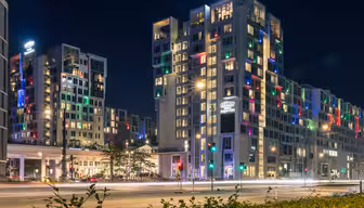 Nattvy av ett modernt hotell med färgglada lampor som lyser från fönstren. Byggnaden är märkt "Tivoli" och omgiven av vägar och grönska. Gatubelysning lyser upp scenen och ljusa färger skapar en livlig atmosfär.