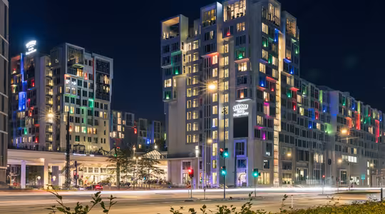 Nattvy av ett modernt hotell med färgglada lampor som lyser från fönstren. Byggnaden är märkt "Tivoli" och omgiven av vägar och grönska. Gatubelysning lyser upp scenen och ljusa färger skapar en livlig atmosfär.