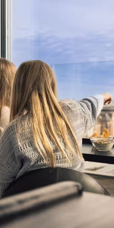 Två kvinnor sitter på ett kafé med stora fönster, med utsikt över en stadsbild. En kvinna pekar ut mot utsikten, som inkluderar stadsbyggnader och en klarblå himmel. De sitter vid ett bord med en liten skål med snacks.