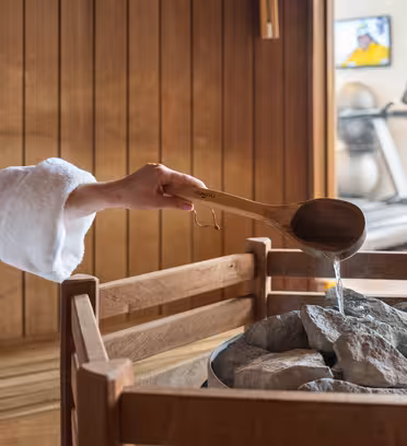 Person i en vit mantel som häller vatten från en träslev på heta stenar i ett basturum i trä. I bakgrunden finns löpband och en TV på väggen.