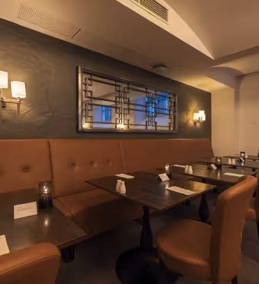 Mysig restauranginteriör med svag belysning. Bruna, dämpade sittplatser kantar väggen, tillsammans med mörka träbord med menyer och små ljus. En dekorativ väggspegel och moderna armaturer ger en stilfull touch. Stort fönster lyser mjukt upp rummet.