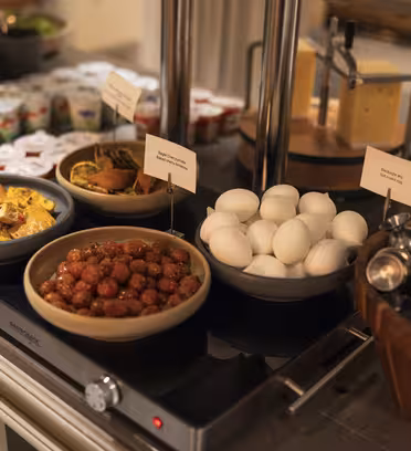 En bufféuppsättning med skålar med äggröra, köttbullar och hårdkokta ägg. I närheten finns koppar yoghurt, ett ostblock och en behållare med små metallmuggar. Den varma maten placeras på en uppvärmd yta.
