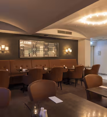 En mysig restauranginredning med bruna läderstolar och träbord. En stor spegel och vägglampor pryder en mörk accentvägg. Mjuk belysning skapar en varm atmosfär, med levande ljus på borden som förstärker den inbjudande atmosfären.