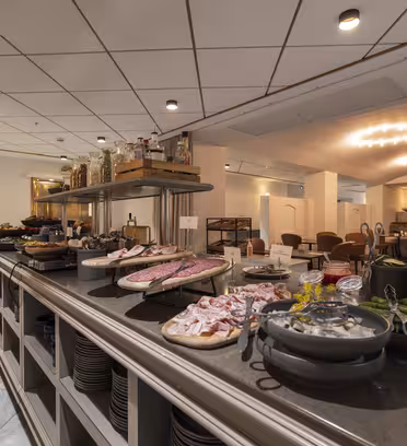 Ett buffébord på en restaurang erbjuder en rad rätter, inklusive skivat kött, ostar, grönsaker och bröd. Matsalen i bakgrunden har snyggt arrangerade bord och stolar under mjuk belysning.
