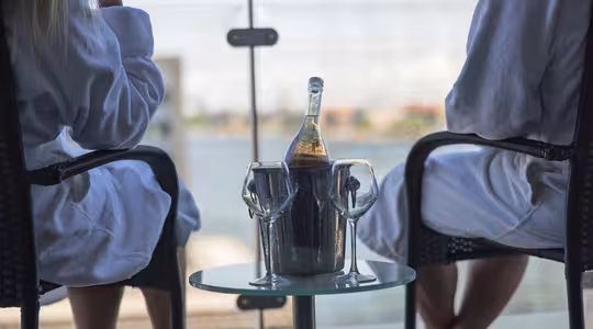 Två personer i vita dräkter sitter på en balkong med utsikt över vattnet. Ett litet bord mellan dem rymmer en vinflaska i en ishink och två glas, som fångar en avkopplande scen med en suddig bakgrund av byggnader och himmel.